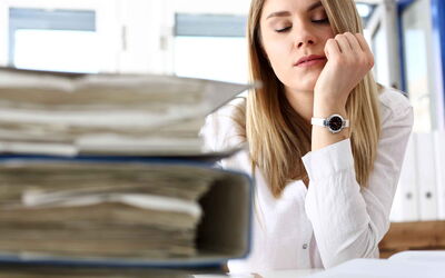 Eine blonde Frau stützt ihren Arm auf einem Schreibtisch auf. Auf dem Tisch liegen viele volle Büroordner. Sie wirkt erschöpft
