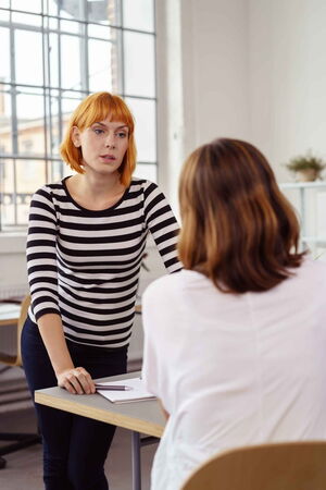 Eine Frau steht vor einer anderen Frau, die mit dem Rücken zu uns sitzt. Die Frau schaut streng. Es scheint einen Konflikt zu geben. Wir helfen Ihnen, Konflikte zu erkennen und zu überwinden und wieder erfolgreich im Team zu arbeiten.