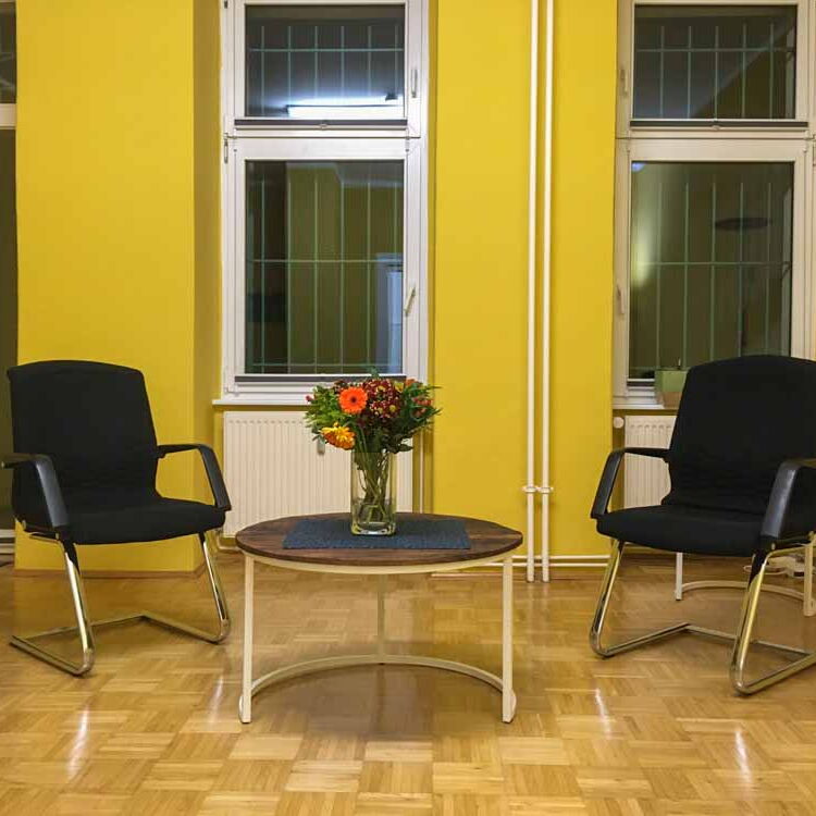 Der große Raum in der Coachingpraxis mit einem runden Tisch und zwei Stühlen vor den Fenstern und der gelben Wand.