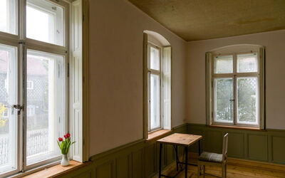 Ein heller, minimalistischer Raum mit großen, minimalistischen Fenstern. Auf der Fensterbank steht eine Vase mit roten Tulpen. In der Ecke des Raumes befindet sich ein Holztisch mit einem schlichten Stuhl, ideal für ruhige Momente oder kreatives Arbeiten.