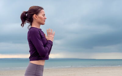 Eine junge Frau steht am Meer. Der Himmel ist wolkig und düster. Sie hat beide Hände vor der Brust verschränkt und die Augen geschlossen. Sie konzentriert sich auf ihren Atem, um zur Ruhe zu kommen.