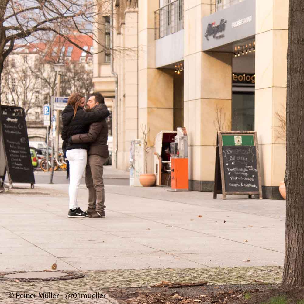 Auf dem Bild ist ein Liebespaar zu sehen. Sie stehen sich gegenüber in dicken Winterjacken und umarmen sich.